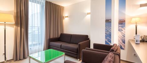 Una habitación de hotel moderna con sofá, sillones, una mesa de vidrio y un escritorio con lámpara.