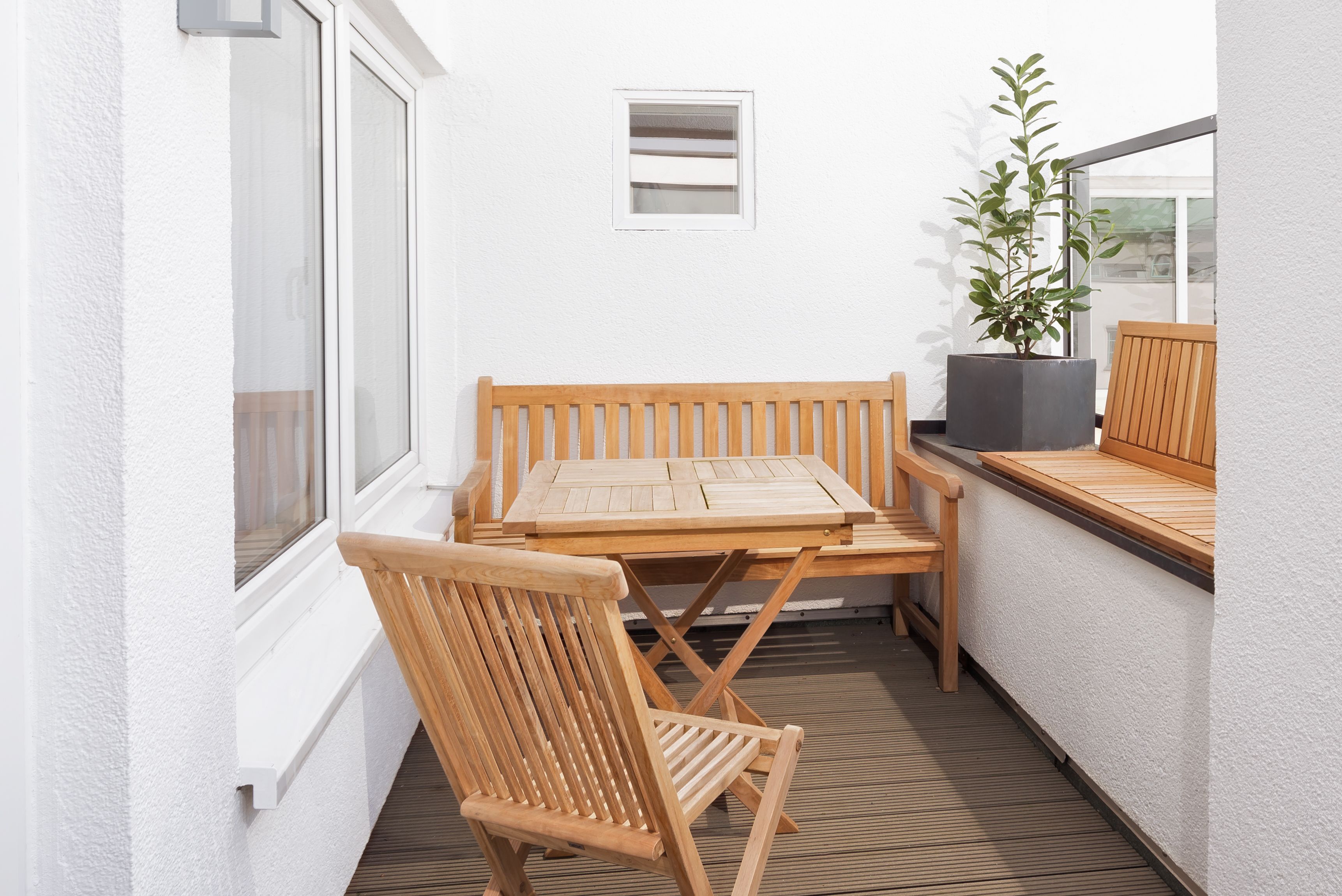 A small outdoor patio with a wooden table and chairs, a potted plant, and a window.