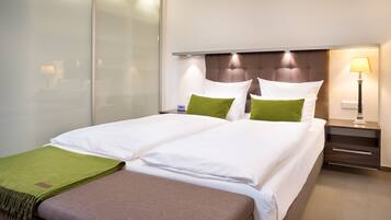 A double bed with white linens and green pillows, a bedside table with a lamp, and a brown bench.