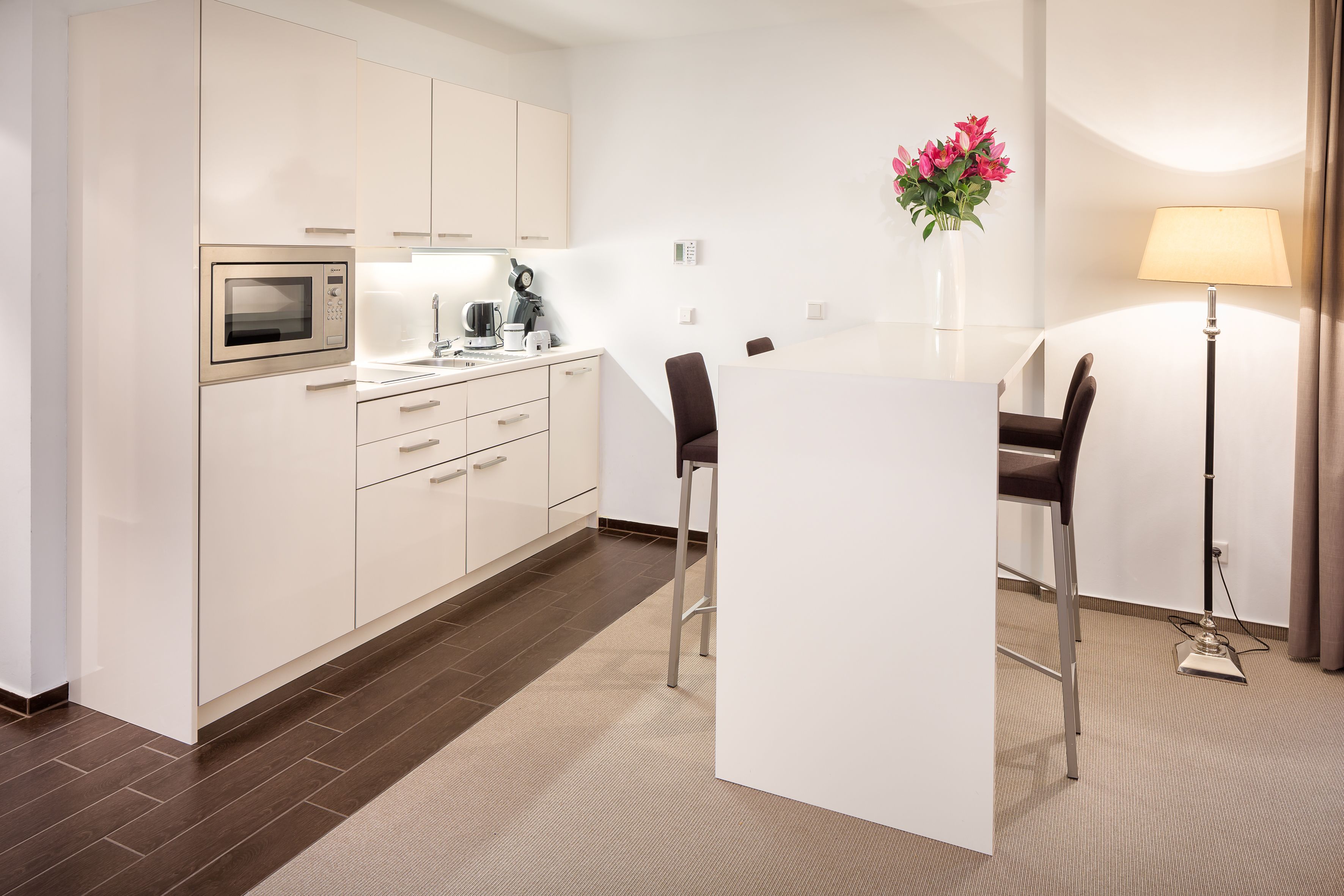A modern kitchen with white cabinets, a microwave, and a coffee maker. There is a dining table with chairs and a vase of flowers on the wall.