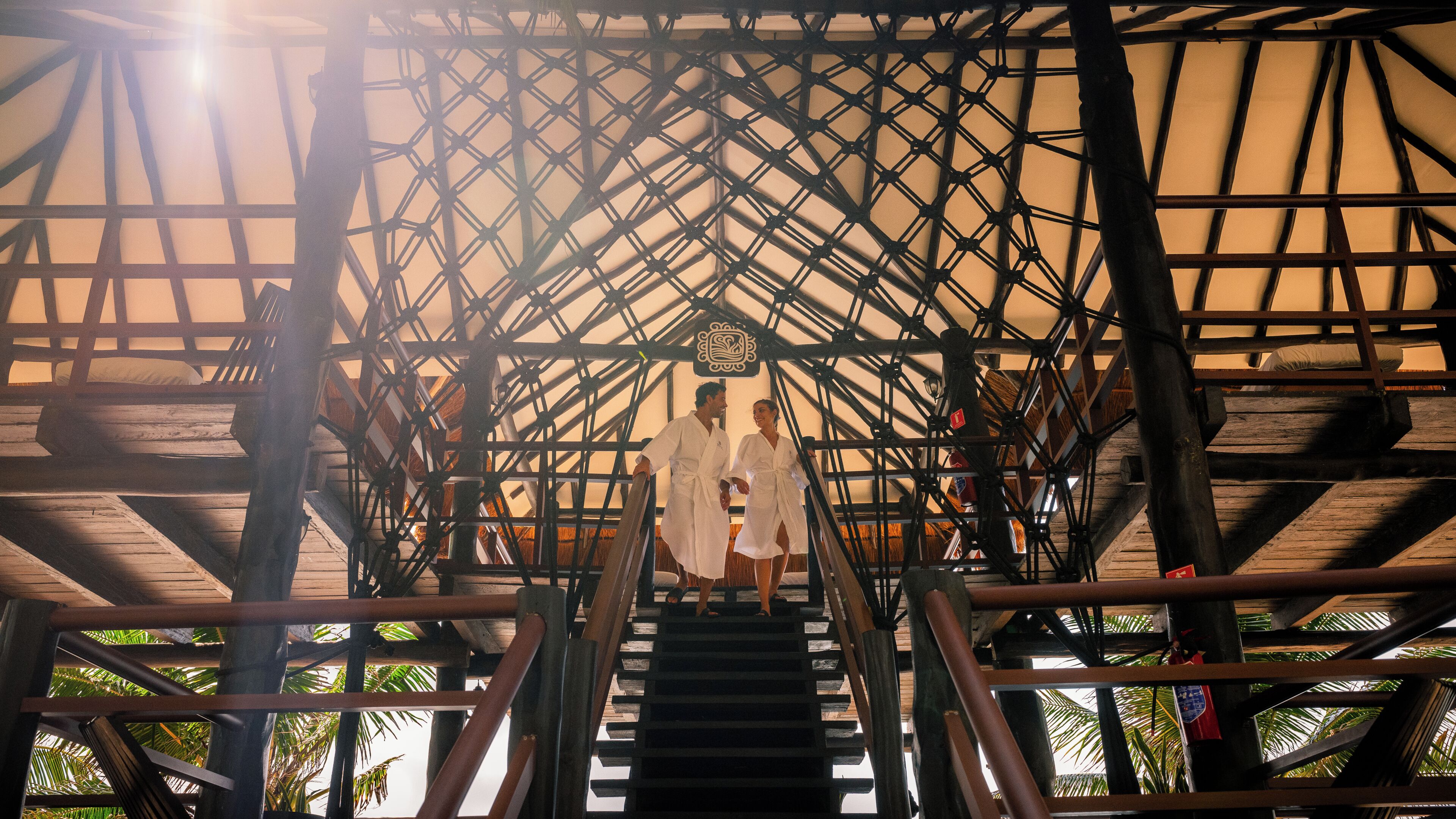 Una escalera de madera con estructura metálica, que lleva a una zona cubierta con vista a palmeras.