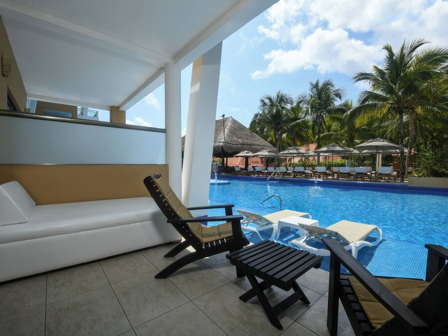 Un espace aménagé au bord de la piscine, avec un canapé blanc, deux chaises longues, une petite table, et une piscine en arrière-plan, agrémentée de parasols et de palmiers.