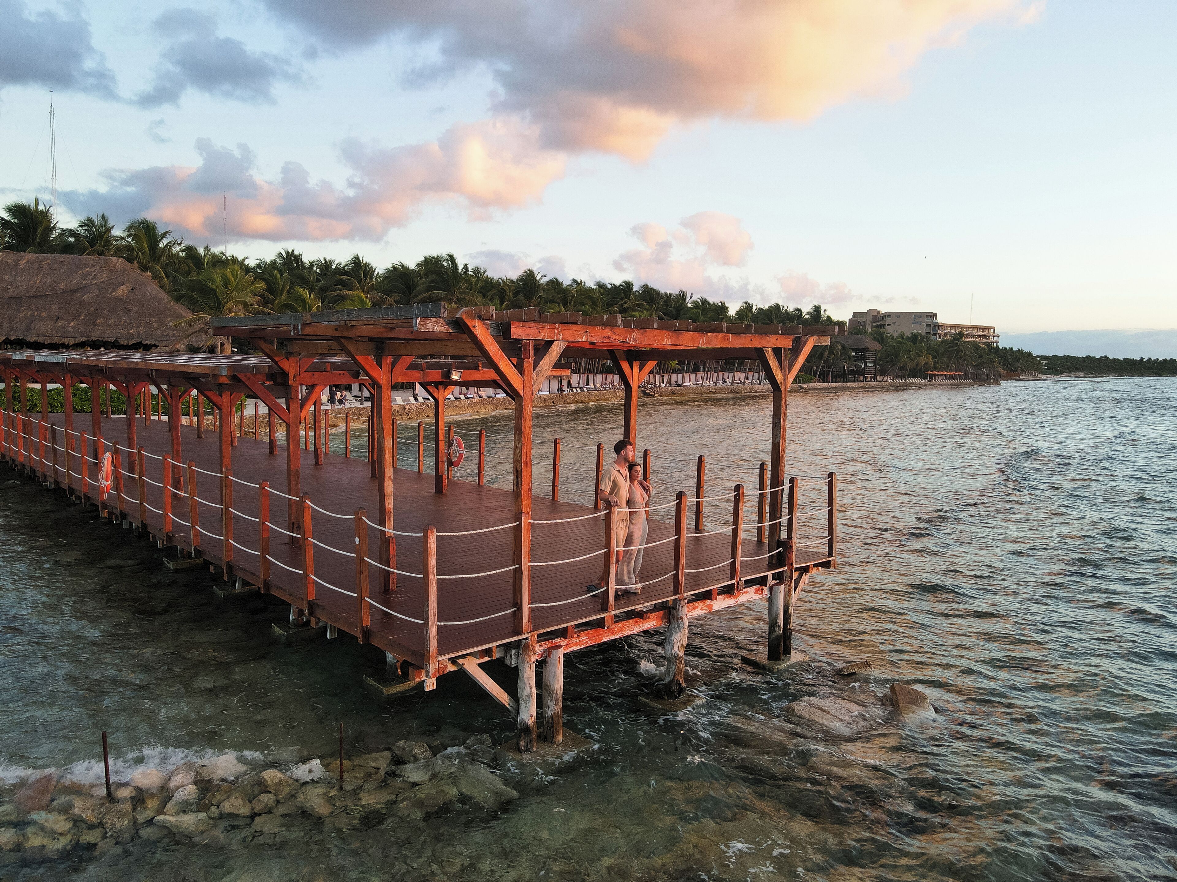 Un muelle de madera que se adentra en una masa de agua, con una estructura de techo de paja en un extremo y un edificio al fondo.