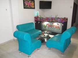 Un hall de hotel con sillones azul verdosos, una pequeña mesa redonda con un jarrón y velas, y un cartel que dice "HOTEL Monarca".