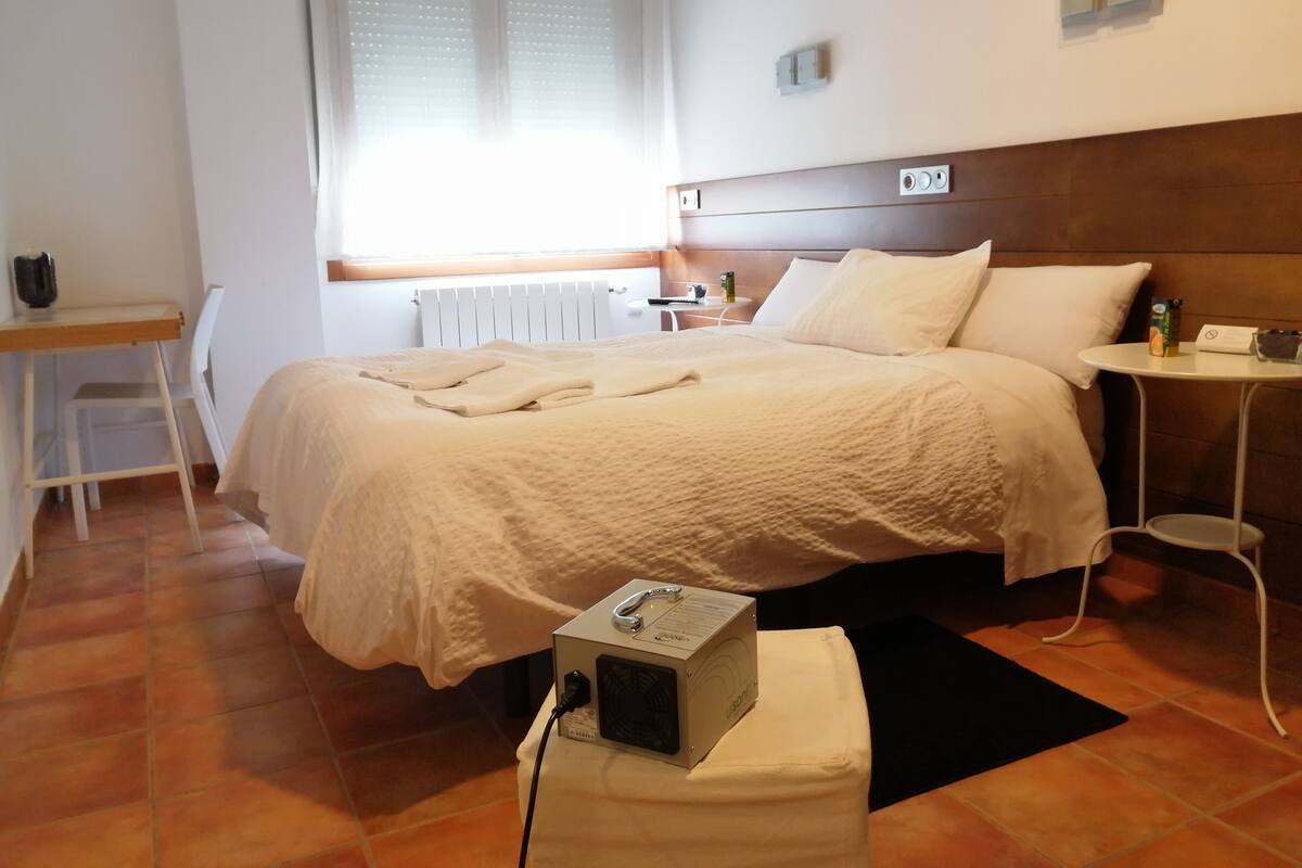 A hotel room with a bed, a desk, a chair, a radiator, a window with blinds, a bedside table, and a power outlet.