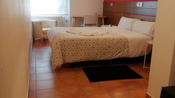Habitación de hotel con cama, escritorio, silla, ventana con persianas, pared de acento roja y lámpara de pie negra.