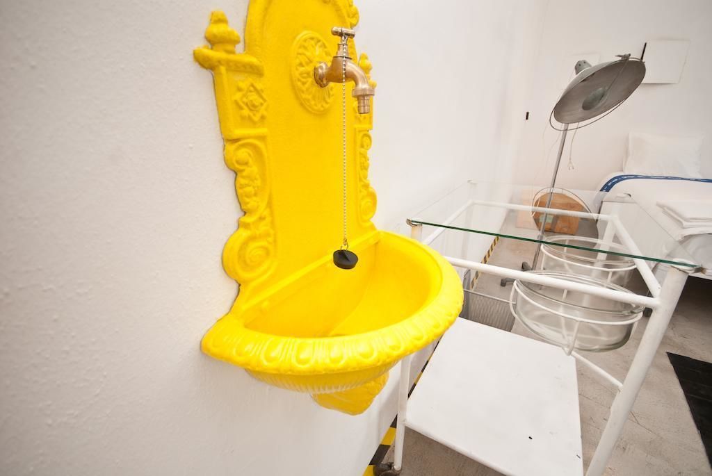 Een geel urinoir aan een witte muur, een witte stoel en een metalen rek met glazen recipiënten.