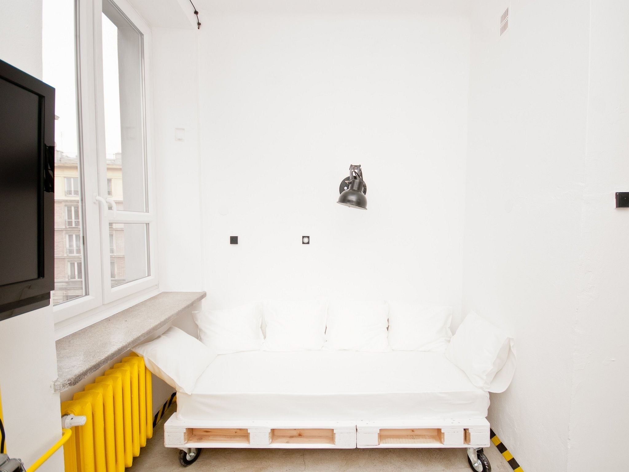 Una habitación pequeña y blanca con una cama individual, un radiador y una ventana.
