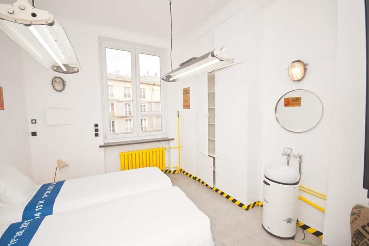 A hotel room with a bed, a window, a radiator, and a white cylindrical object on the floor.