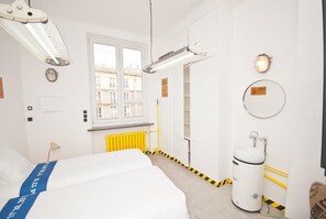 A hotel room with a bed, a window, a radiator, and a white cylindrical object on the floor. - LoftHotel Przychodnia (Warsaw)