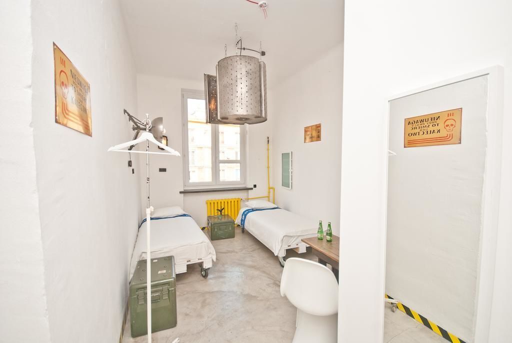 Habitación pequeña y limpia con dos camas, un radiador, una lámpara colgante y una puerta con un cartel de advertencia.