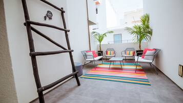 A small, enclosed patio area with a wooden ladder, two chairs with colorful cushions, a striped rug, and two potted plants.