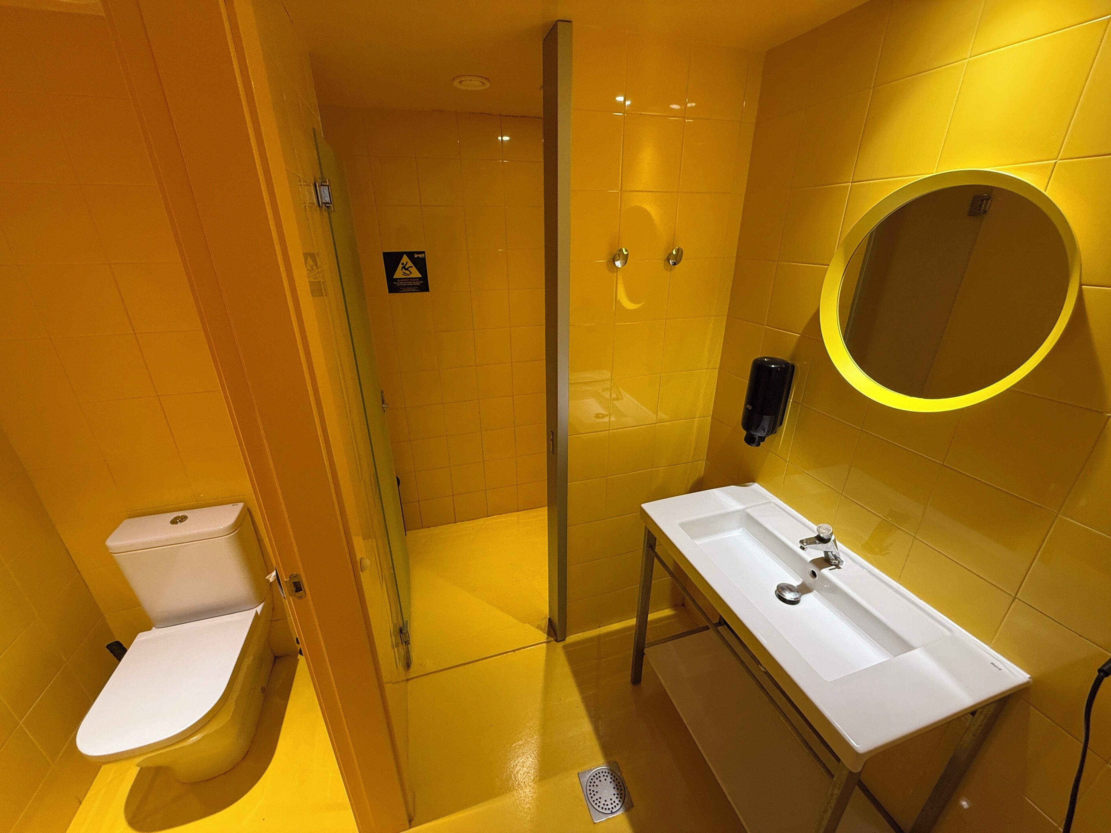 Une salle de bain moderne avec un mur recouvert de carreaux jaunes, des toilettes blanches, une douche en verre et un lavabo blanc avec un miroir.