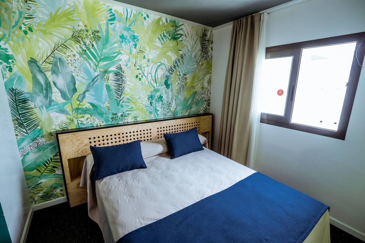 Une chambre d’hôtel avec un lit, une fenêtre avec des rideaux et un mur orné d’un motif de feuilles tropicales.