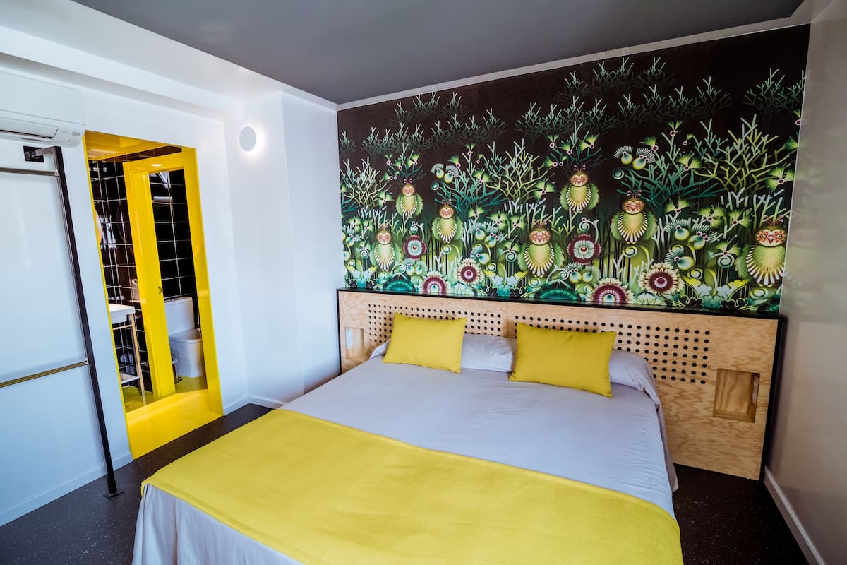 Une chambre à coucher avec un lit, une décoration aux couleurs jaune et blanc, une tête de lit ornée d’une fresque florale et une armoire avec une porte jaune.