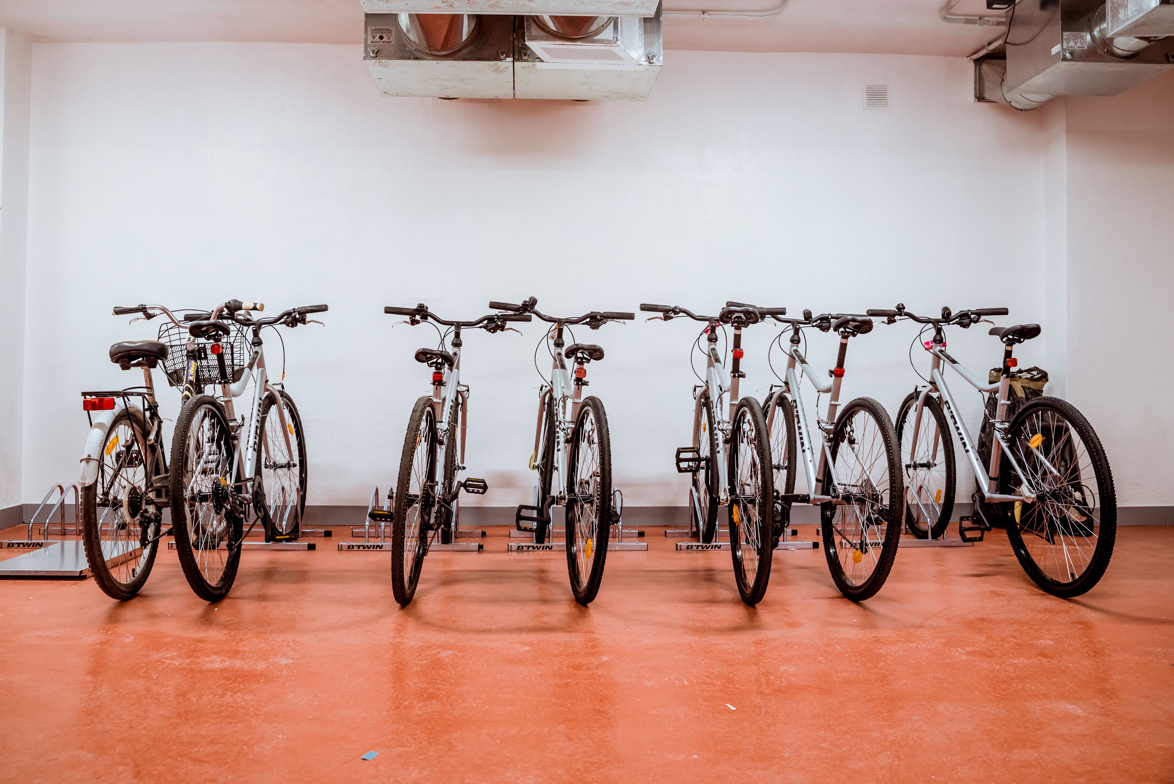 A row of bicycles parked indoors with a plain white wall and a red floor.