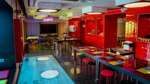A modern restaurant with a vibrant interior, featuring a long communal table with stools, red and yellow checkered flooring, and overhead lighting.