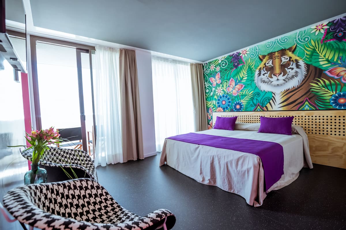Une chambre d’hôtel avec un lit, un fauteuil à motif pied-de-poule, une grande fenêtre avec des rideaux et une fresque murale représentant un tigre aux couleurs vives.