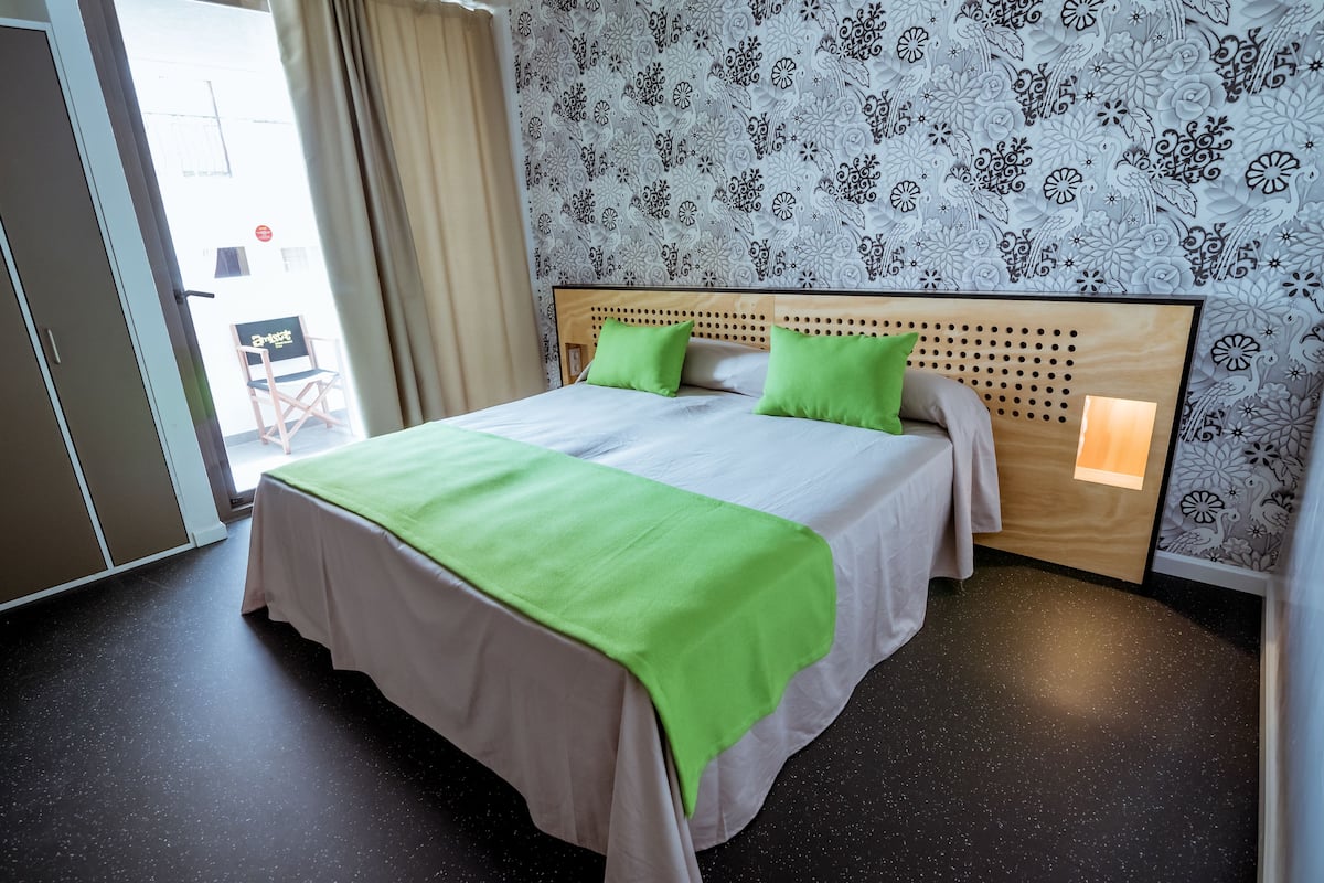 Une chambre d’hôtel avec un lit, des oreillers verts, une tête de lit en bois, un papier peint à motifs et une porte donnant sur l’extérieur.