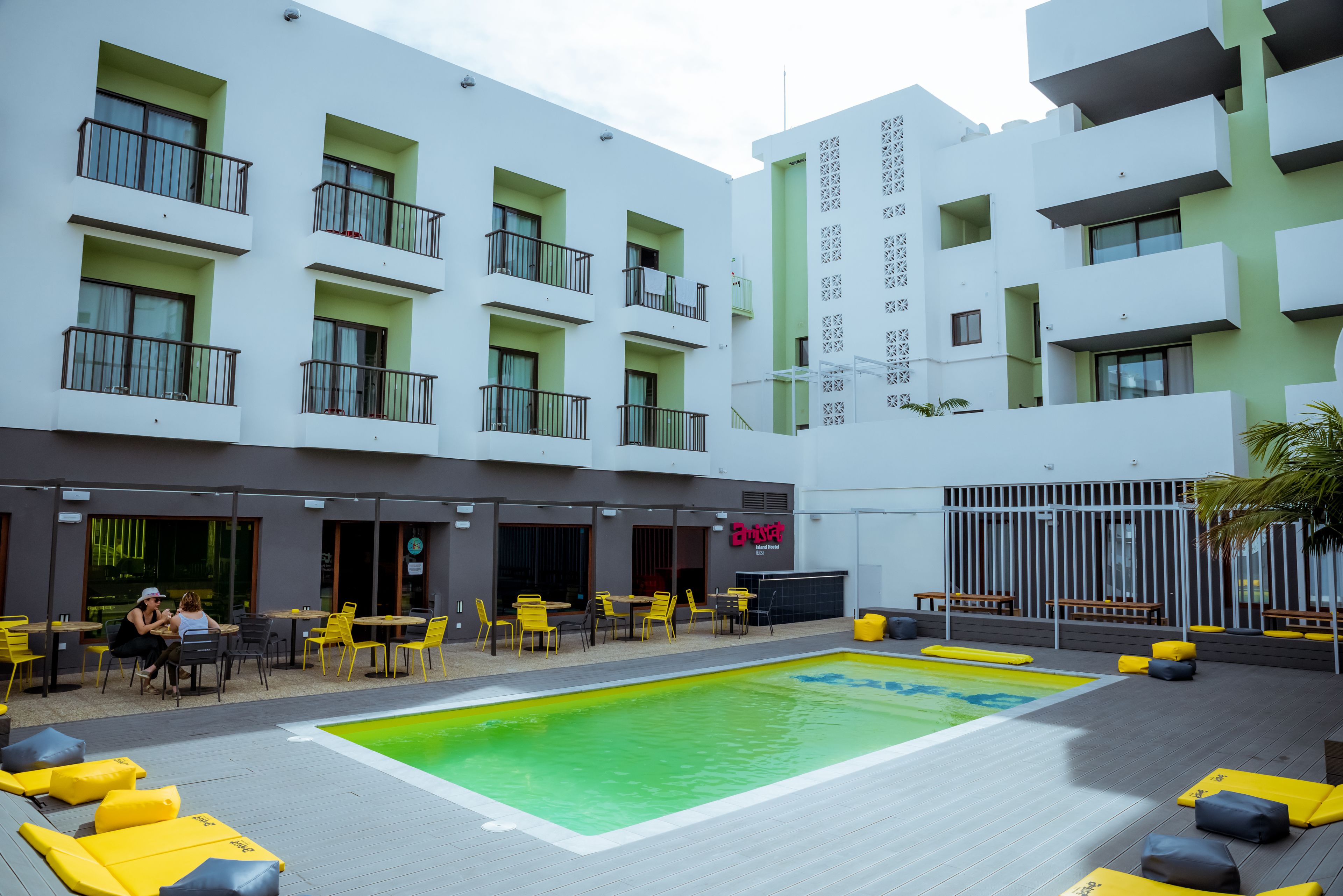 A rooftop pool area with yellow lounge chairs, a dining area with tables and chairs, and a building with balconies and a sign that reads "Café".