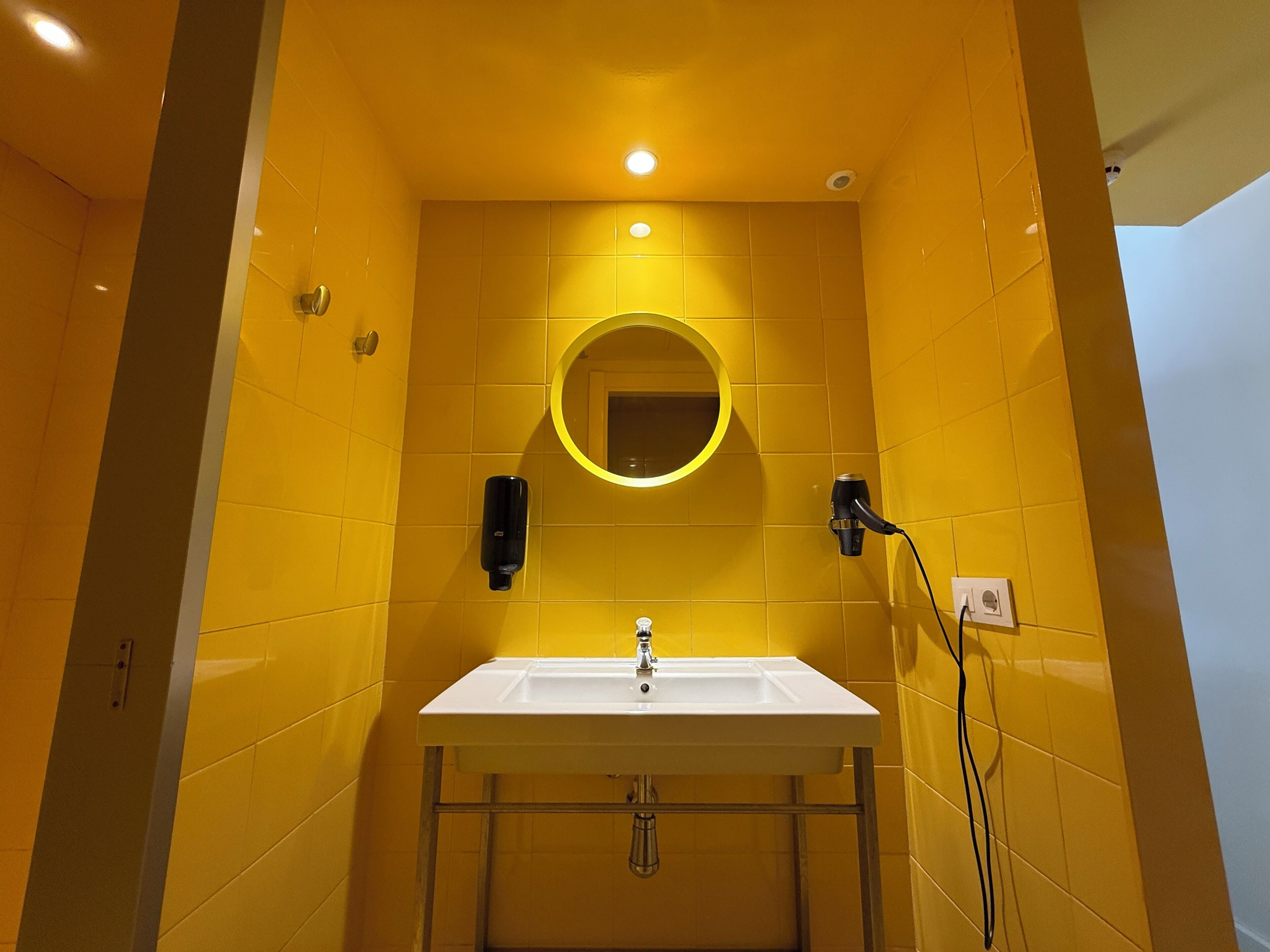 Un baño con paredes amarillas, un lavabo blanco, un espejo redondo y un secador de pelo enchufado.