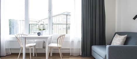 Una sala de estar moderna con un sofá gris, una mesa de comedor con dos sillas y vistas a edificios a través de amplios ventanales.