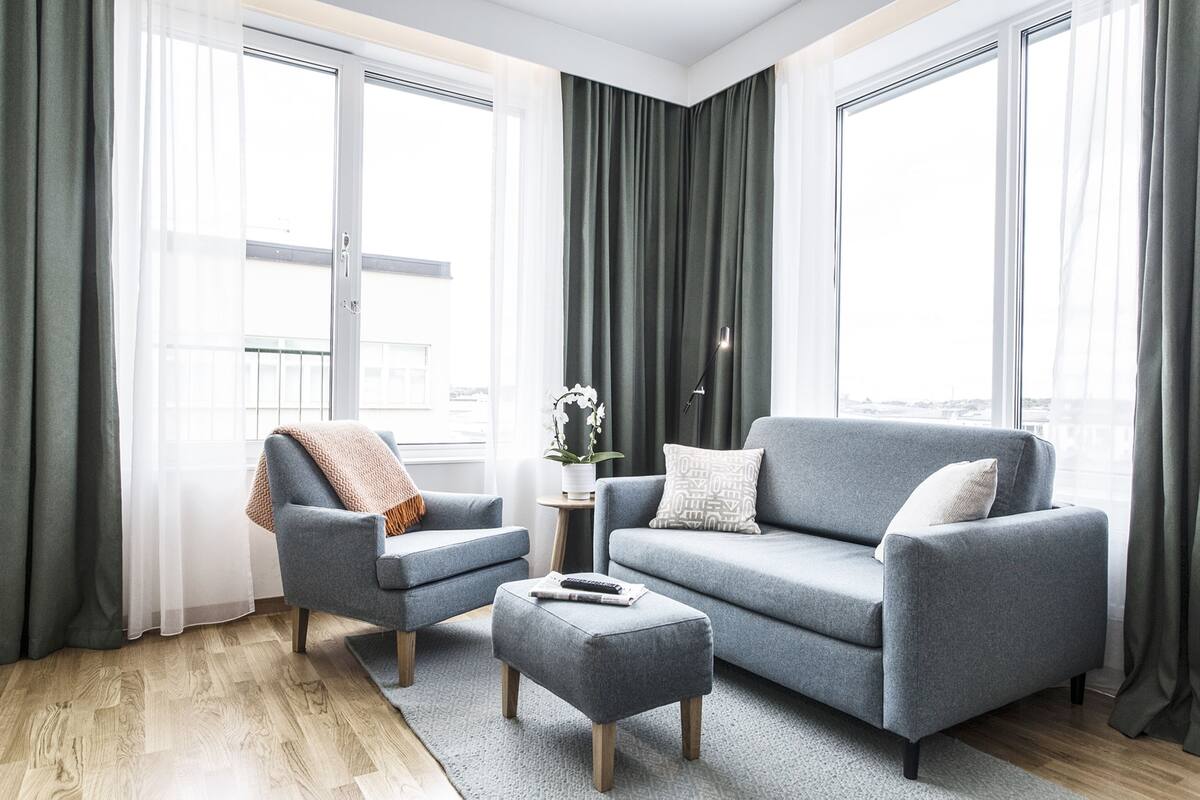Een moderne woonkamer met een grijze bank, een bijpassende fauteuil en een poef. Er staat een klein bijzettafeltje met een plant en een lamp.