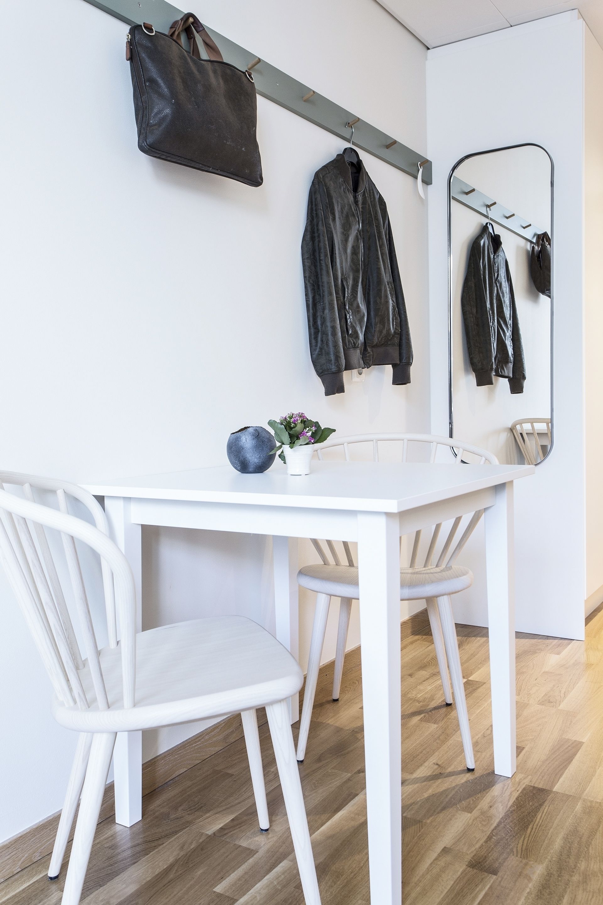 Een witte eettafel met twee stoelen, een hangende lederen tas en een spiegel aan de muur.