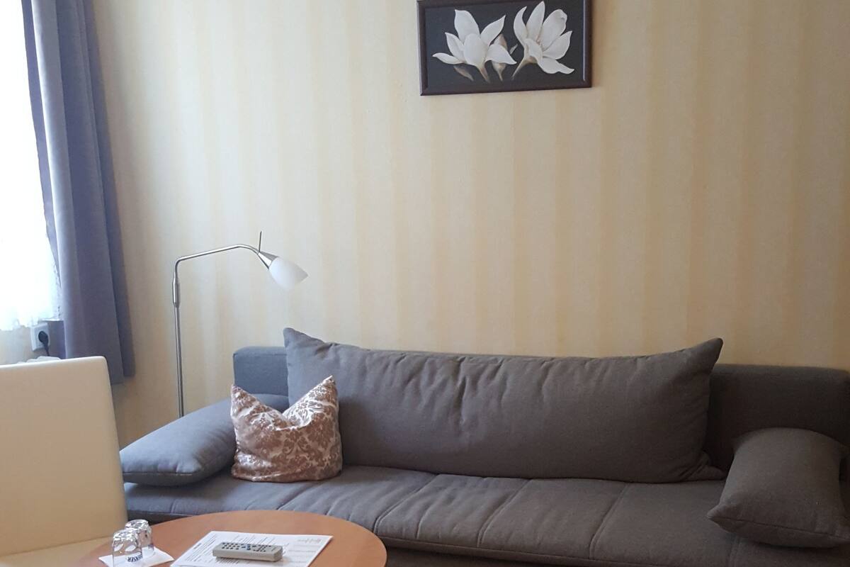 A hotel room with a bed, a gray sofa, a small round table with a remote control, a lamp, and a framed picture on the wall.