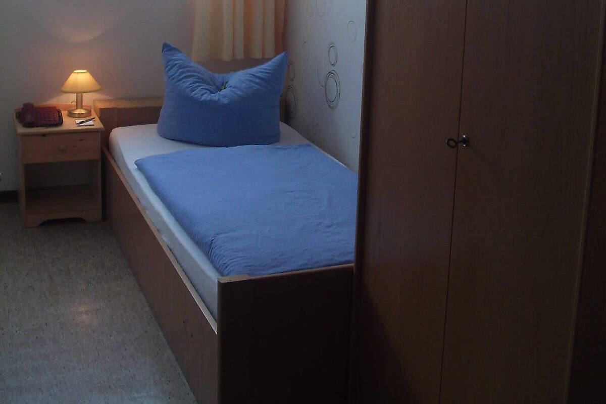 A small, neatly arranged bedroom with a single bed, a nightstand with a lamp, a wardrobe, and a window with curtains.