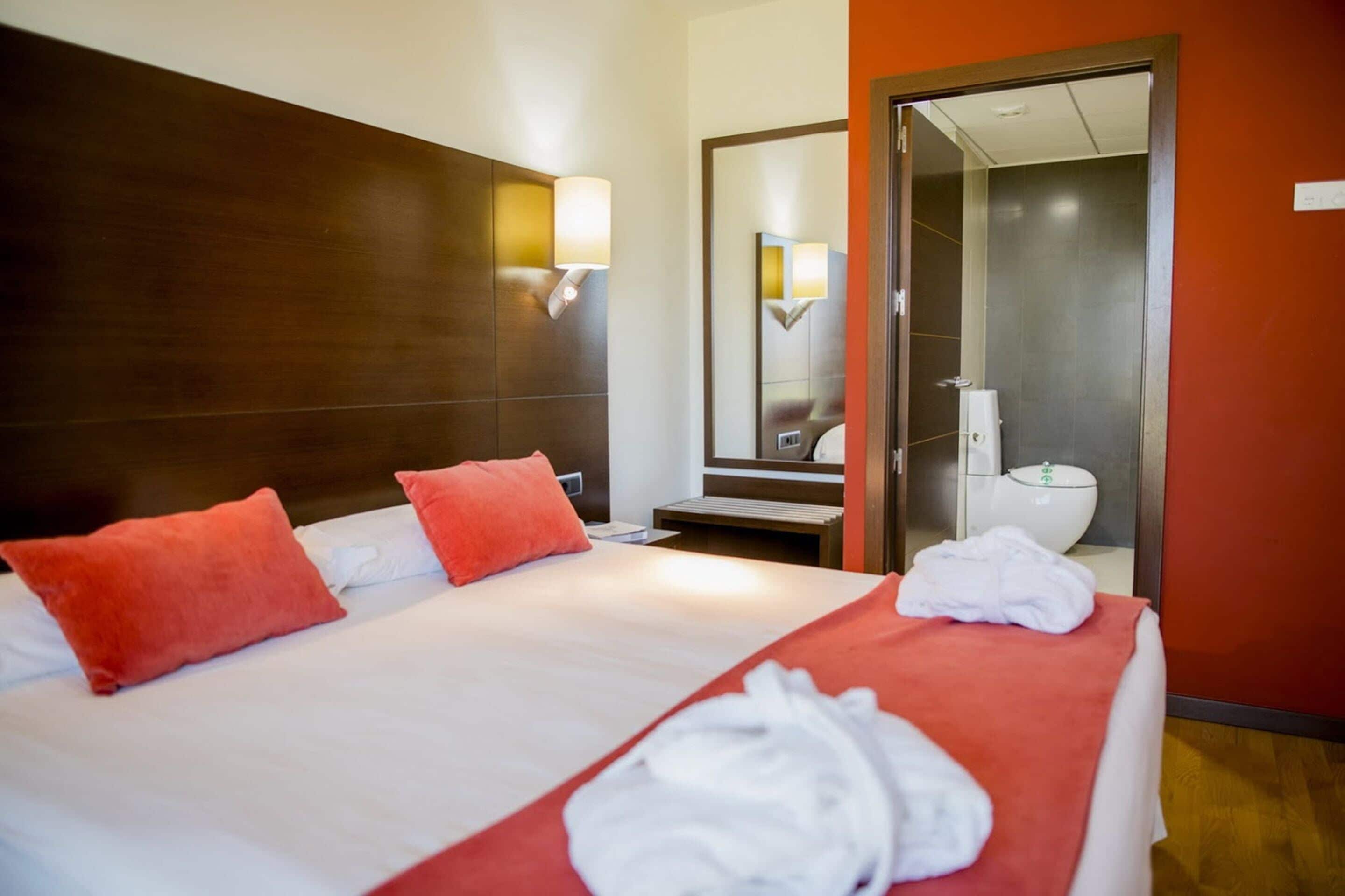 Une chambre d’hôtel avec un grand lit, deux oreillers rouges, une couverture rouge et une serviette blanche posée sur le lit. On peut voir une tête de lit en bois, un miroir et une salle de bain à travers une porte ouverte.