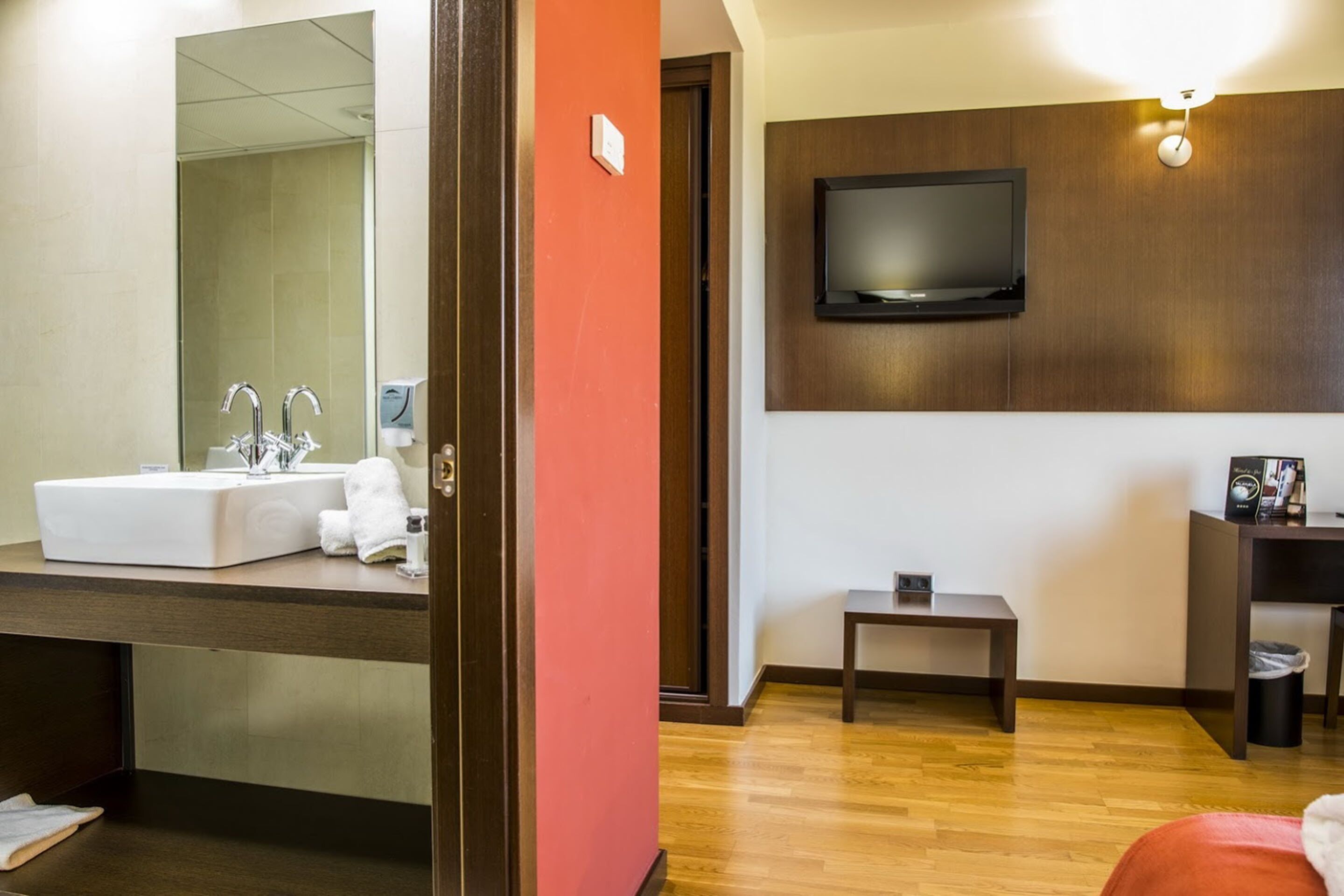 Ein Hotelzimmer mit einem großen Spiegel, einem weißen Waschbecken, Holzboden, einem kleinen Tisch, einem an der Wand montierten Fernseher und einer roten Akzentwand.