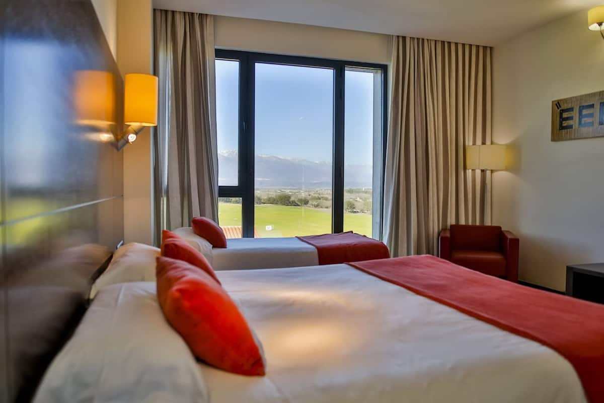 Een hotelkamer met een groot raam, een bed met rode kussens, een stoel en een lamp.