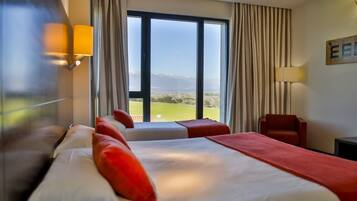 A hotel room with a large window, a bed with red pillows, a chair, and a lamp.