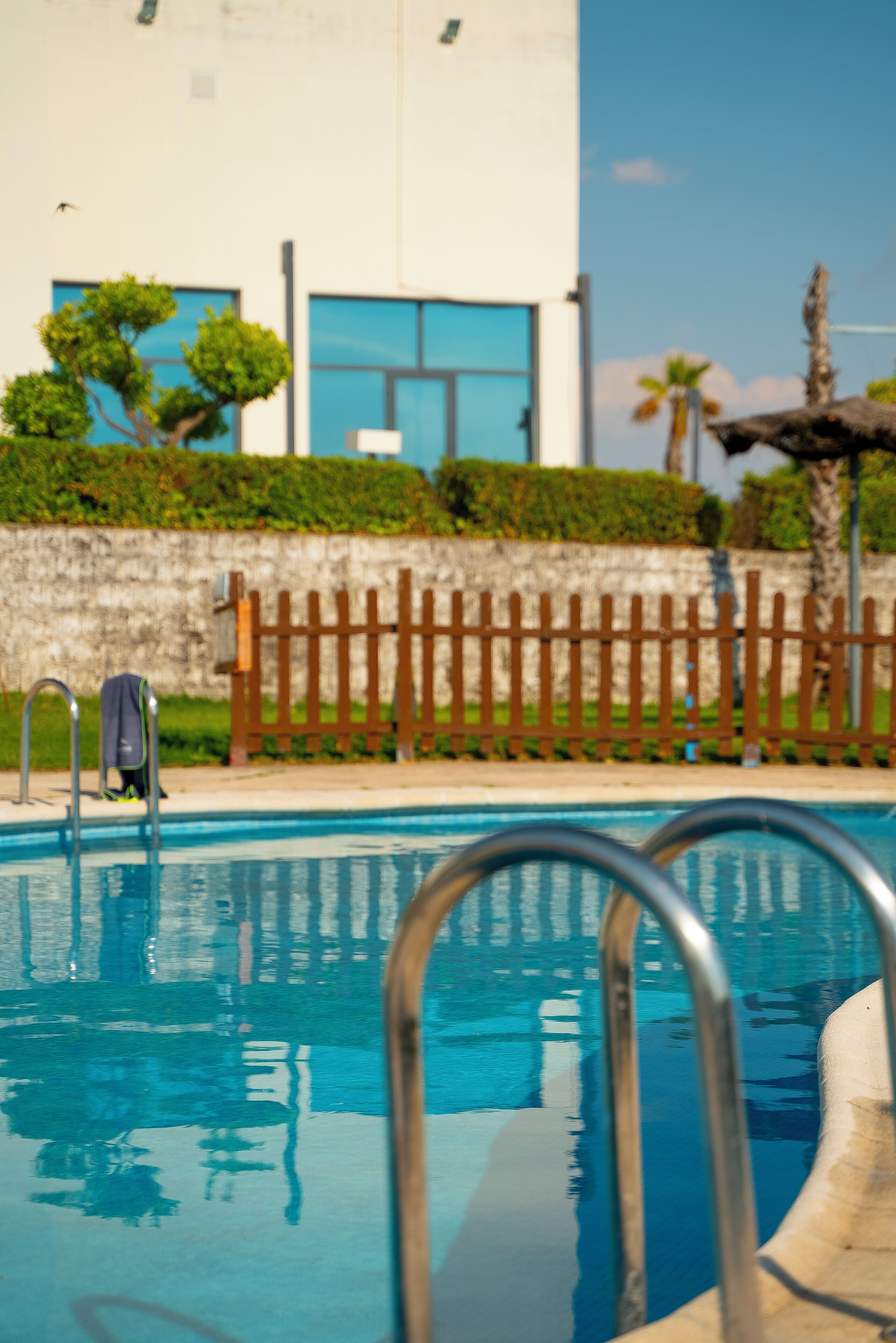 Una piscina con barandillas metálicas, una valla de madera y un edificio con ventanas amplias al fondo.