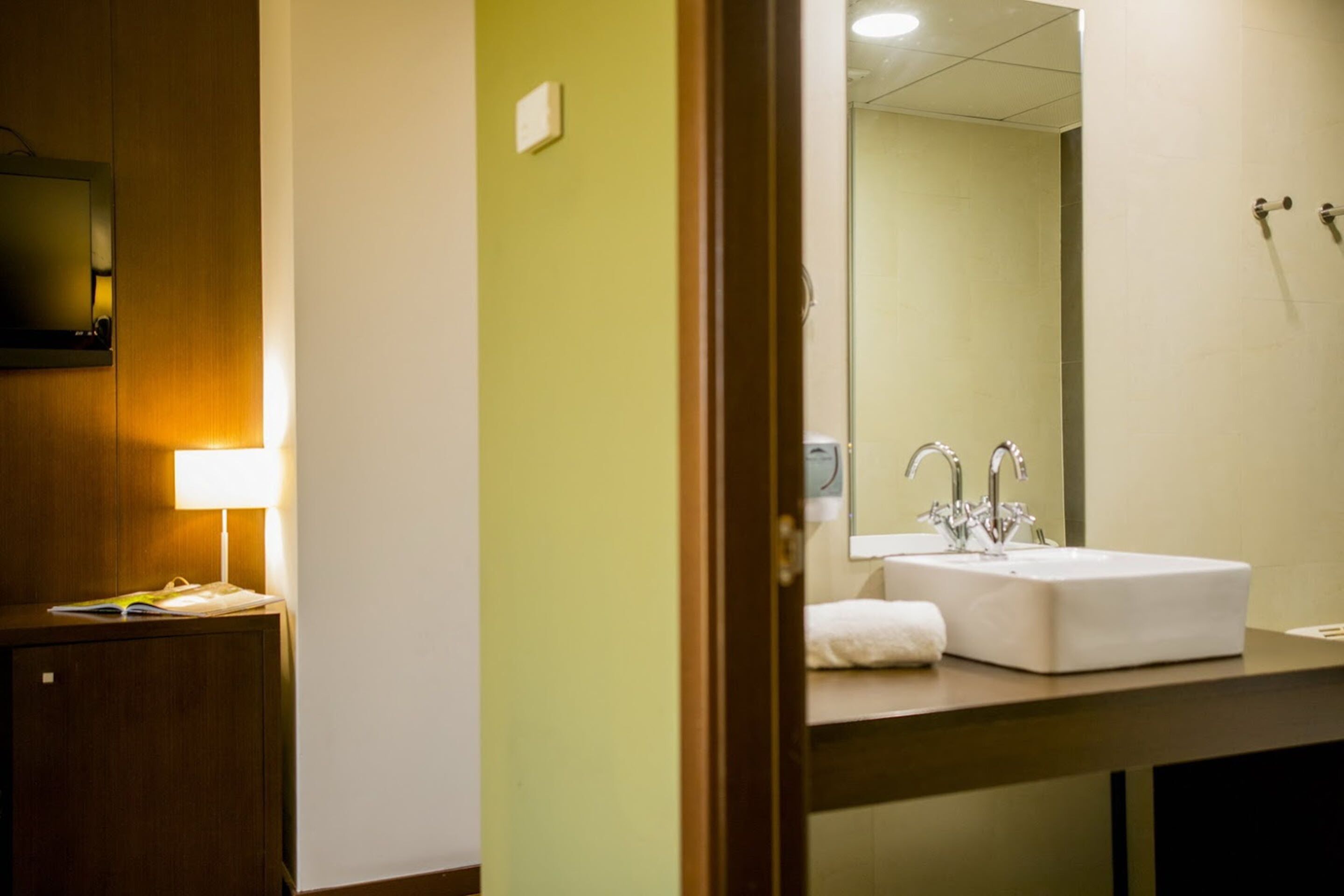 A hotel bathroom with a sink, mirror, and a wooden cabinet with a lamp.