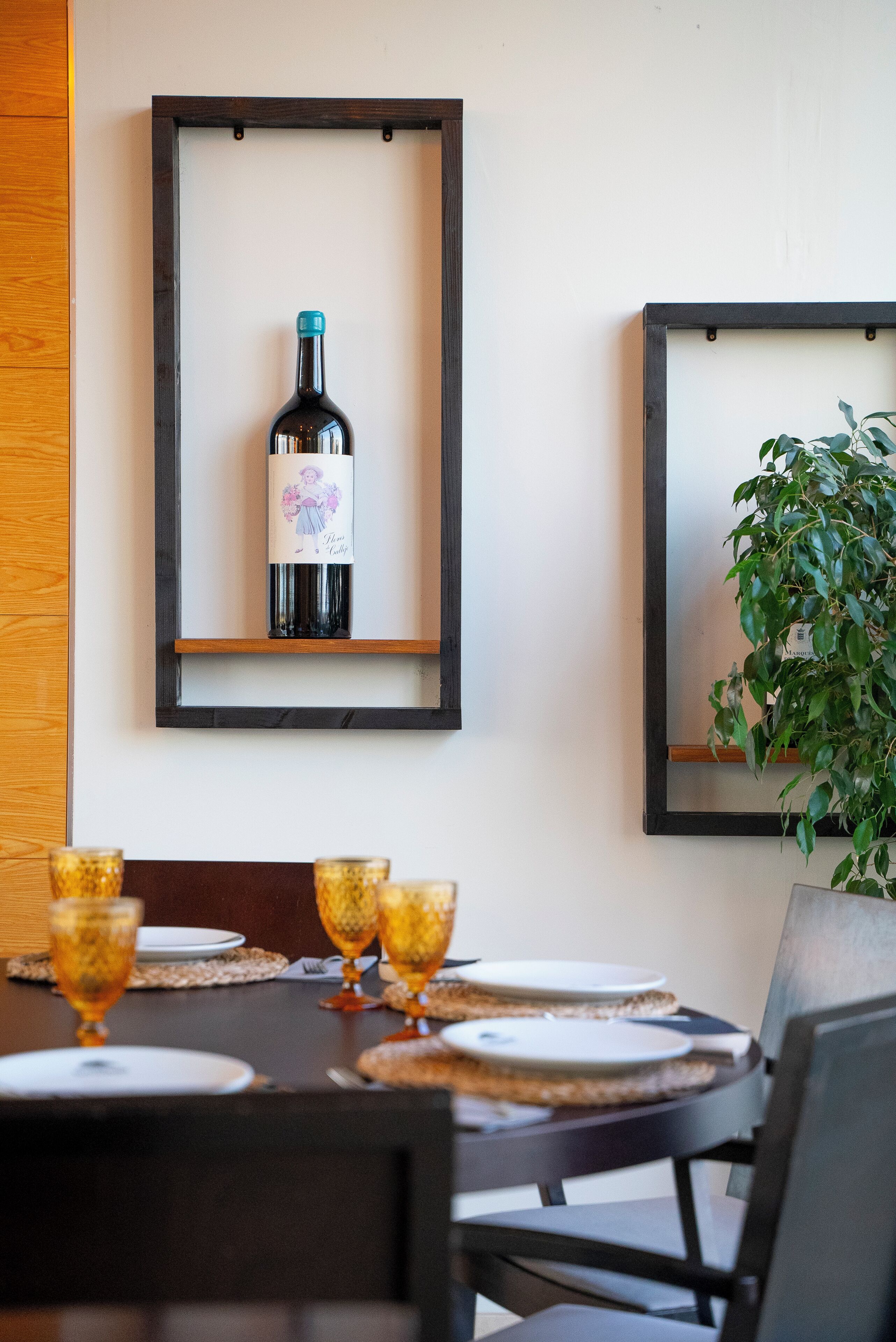Una mesa de comedor con una botella de vino, copas, una obra de arte enmarcada y una planta en maceta.
