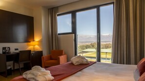 Habitación de hotel con una cama grande, un escritorio, una silla naranja y vistas a un paisaje montañoso a través de ventanas amplias.