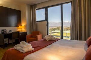 Habitación de hotel con una cama grande, un escritorio, una silla naranja y vistas a un paisaje montañoso a través de ventanas amplias.