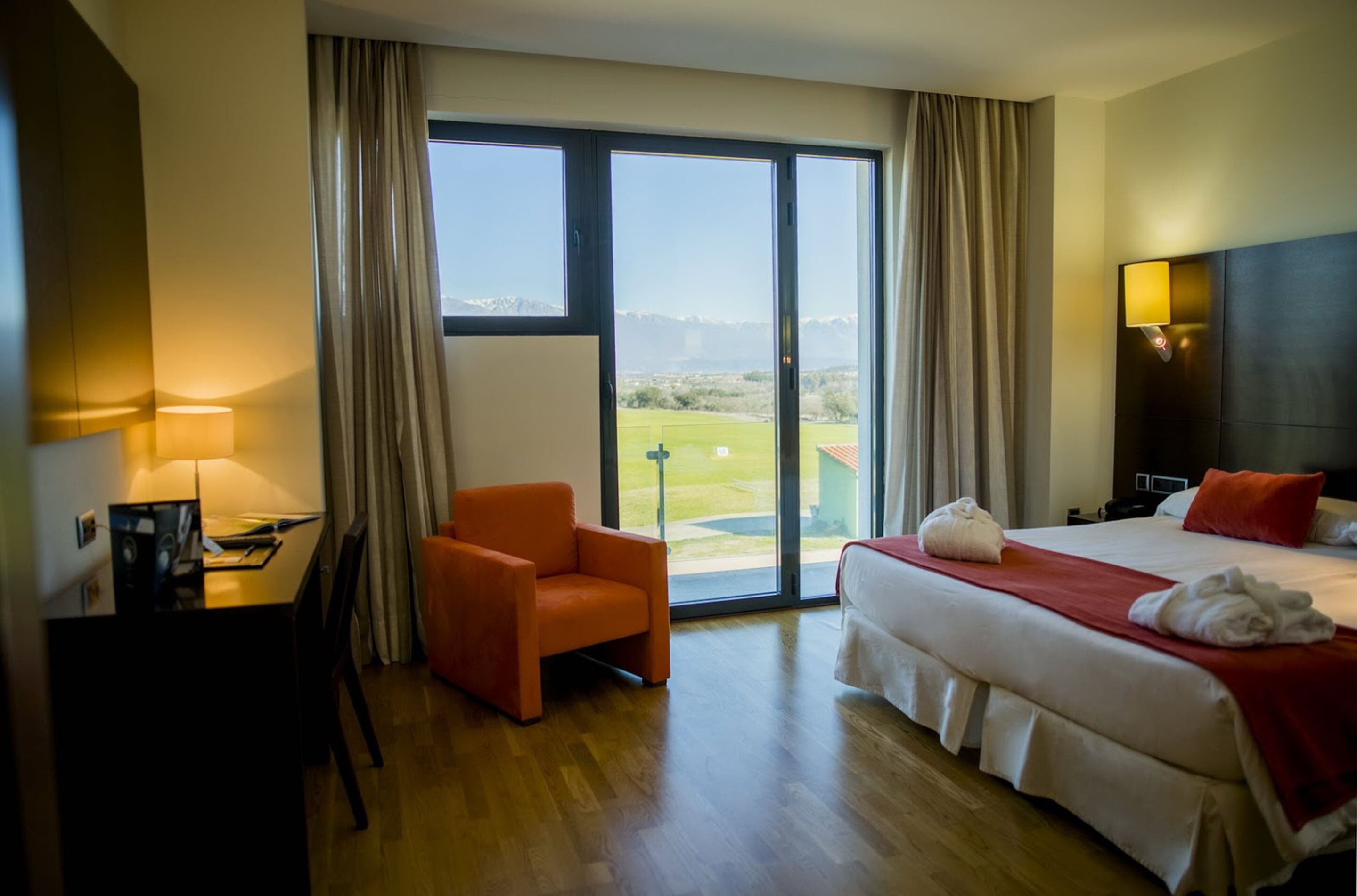 Habitación de hotel con cama, escritorio, silla y un ventanal con vistas a un paisaje verde.