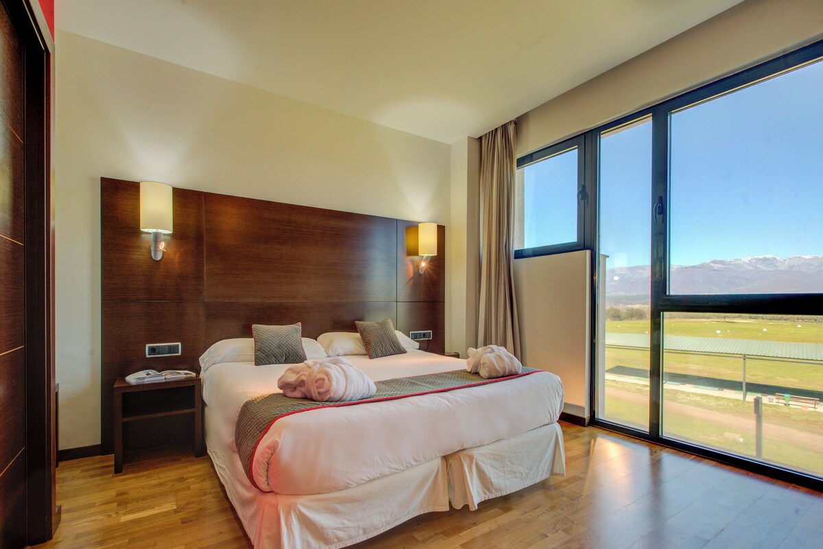 Amplia habitación de hotel con una cama grande, una mesita de noche y vistas a un paisaje verde a través de amplias ventanas.