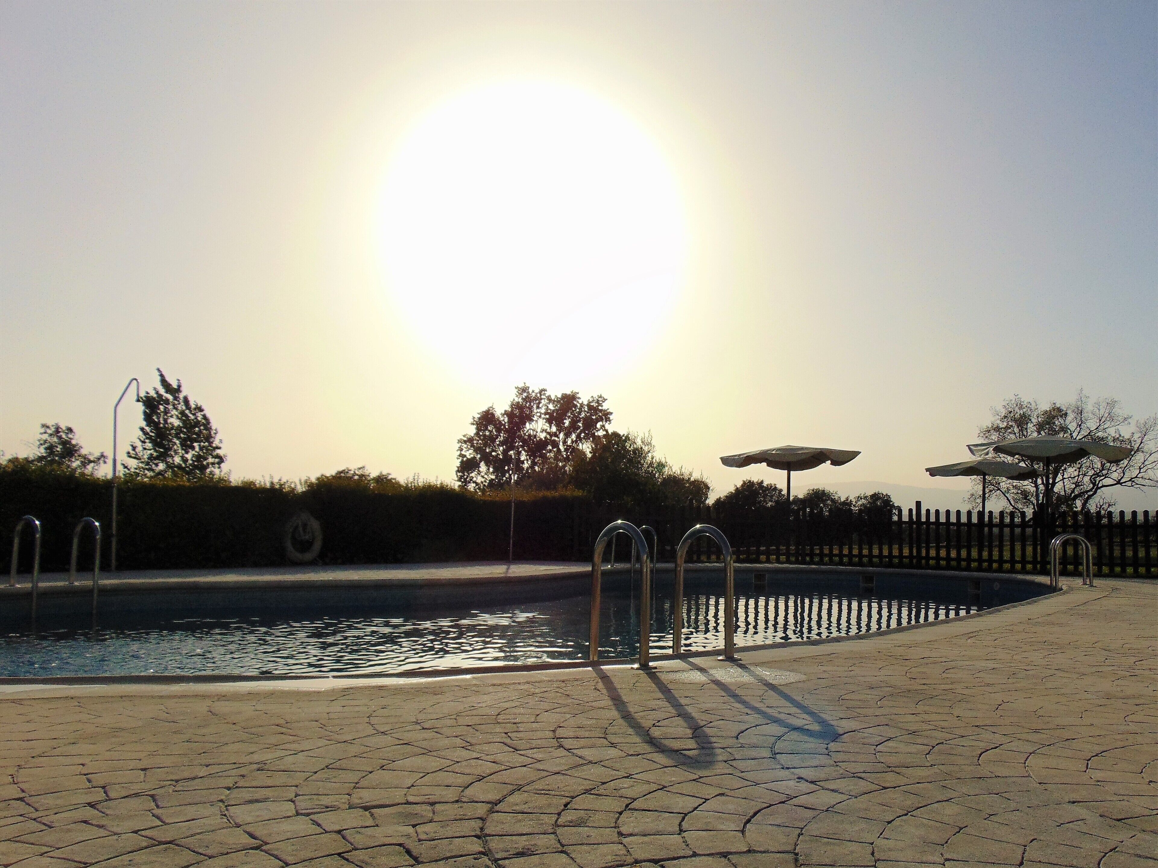 A swimming pool with a sunlit sky, metal pool ladders, and a paved walkway.
