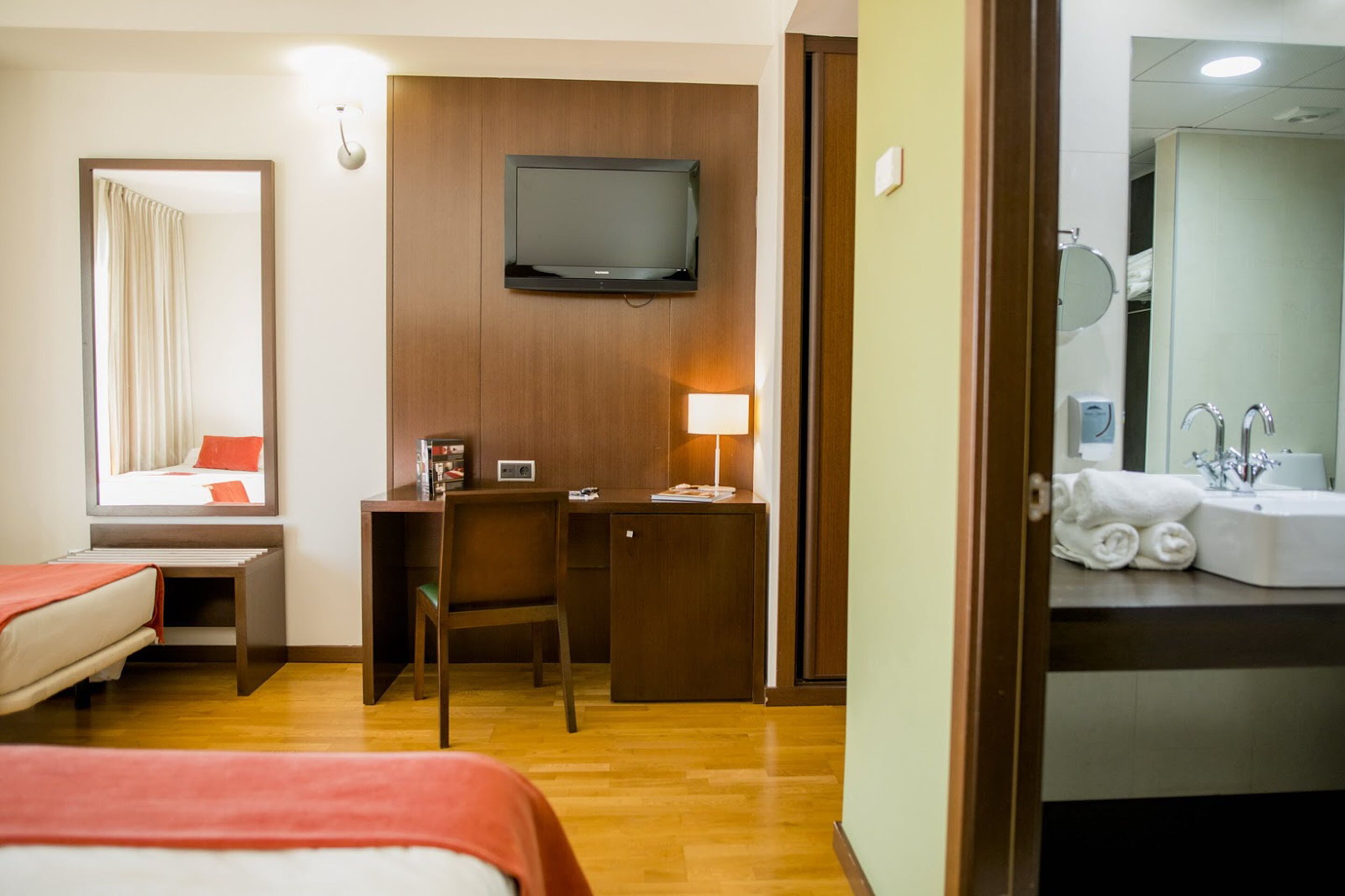 A hotel room with a bed, a desk, a TV, and a bathroom with a sink and mirror.