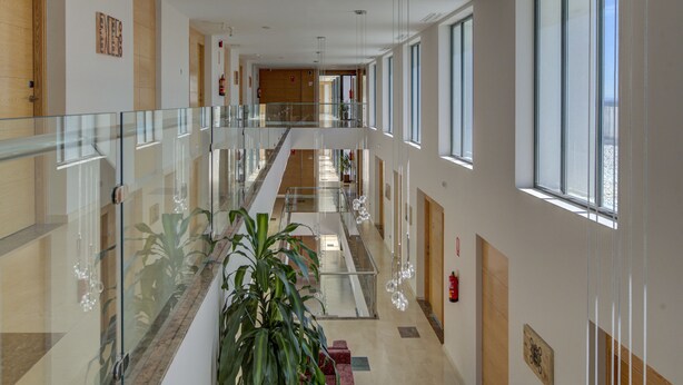 Un pasillo moderno de hotel con barandillas de vidrio, una escalera central y ventanas amplias que permiten la entrada de luz natural.