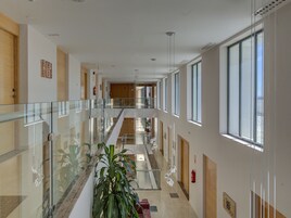 Un pasillo moderno de hotel con barandillas de vidrio, una escalera central y ventanas amplias que permiten la entrada de luz natural.