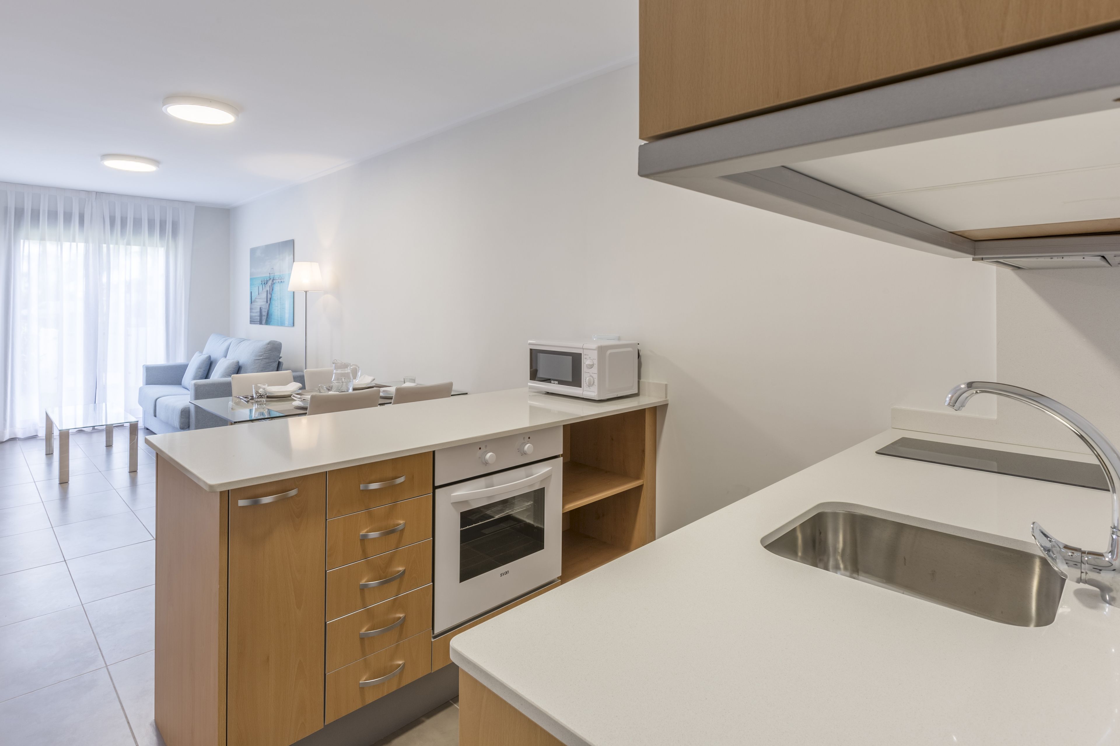 Cocina moderna con encimeras blancas, horno empotrado, microondas y pileta de acero inoxidable.