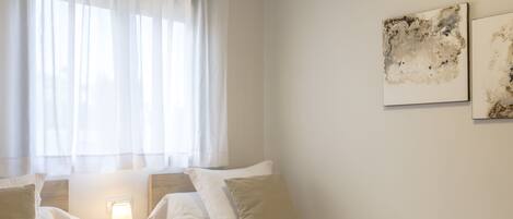 A hotel room with two single beds, a bedside table with a lamp, and a window with sheer curtains.