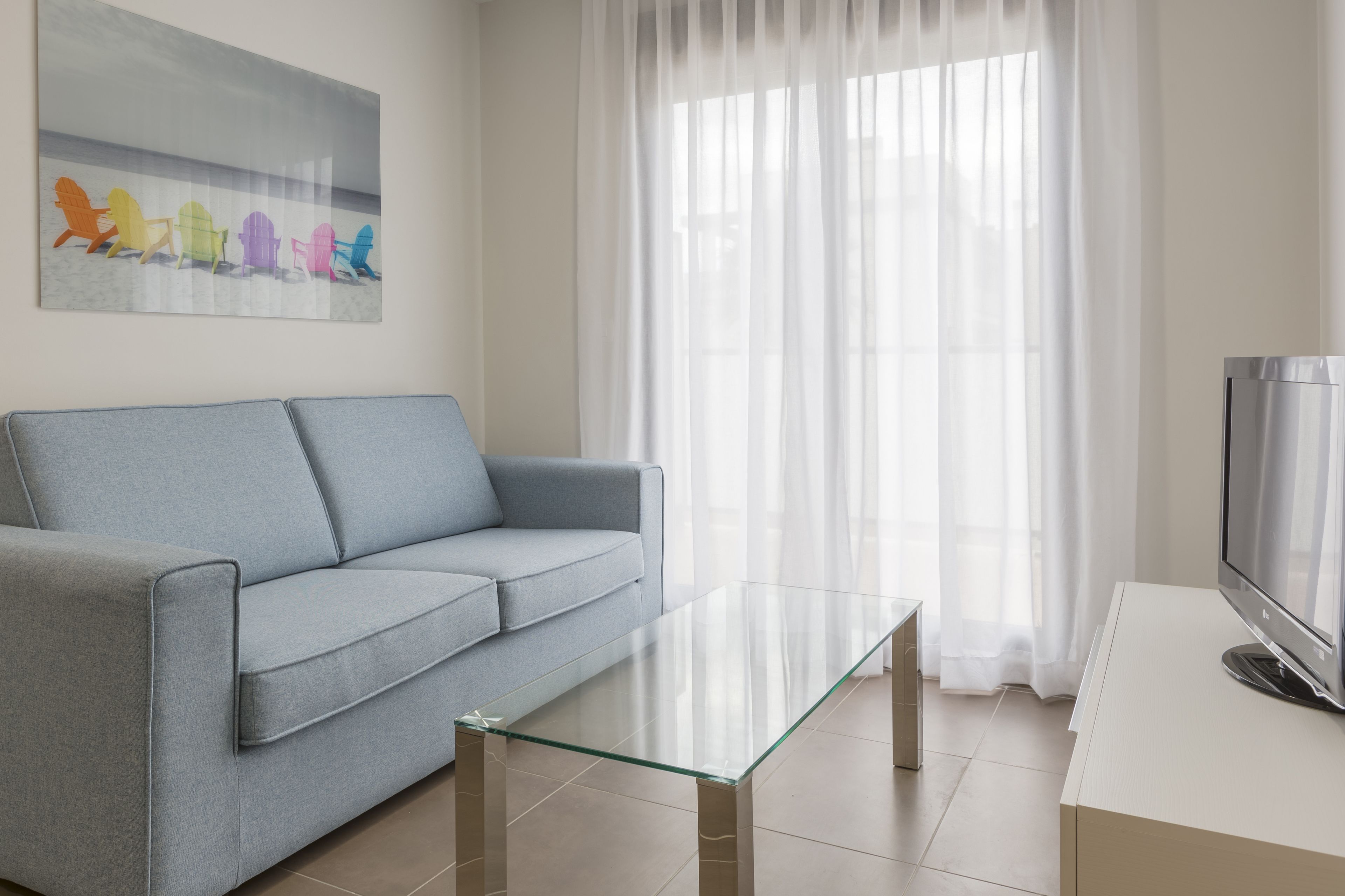 Un salon moderne avec un canapé gris, une table basse en verre, un téléviseur à écran plat posé sur un meuble et une grande fenêtre ornée de rideaux légers.