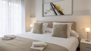 Ein ordentlich bezogenes Bett mit einer beigen Tagesdecke und Kissen, ein Nachttisch mit einer Lampe und ein Bild an der Wand.