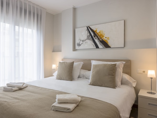 Una cama bien hecha con una colcha y almohadas color beige, una mesita de noche con una lámpara, y un cuadro en la pared.